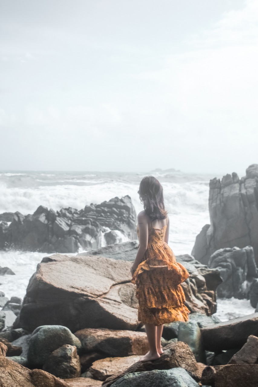 young-girl-in-dress-standing-on-large-rocks-watching-waves