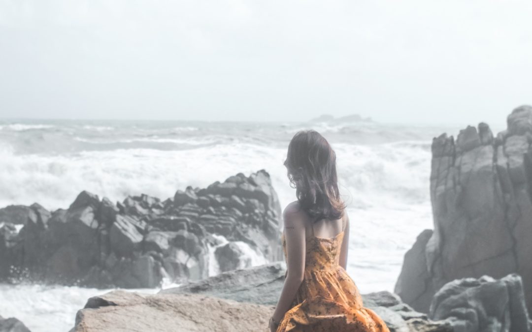 young-girl-in-dress-standing-on-large-rocks-watching-waves.jpg